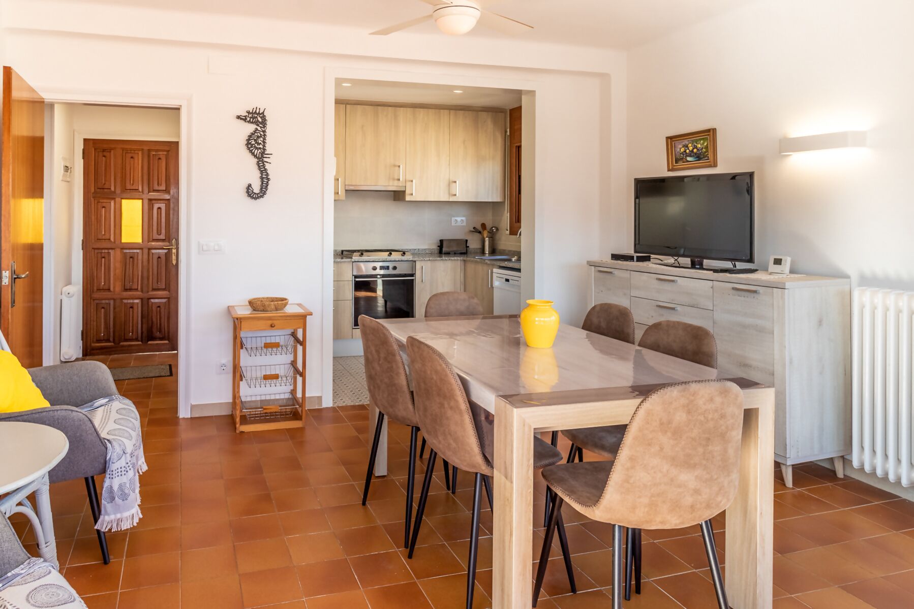 A dining area with a table and chairs, a kitchen in the background, and a TV on a cabinet.