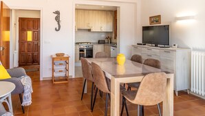 Zona pranzo con tavolo e sedie, cucina sullo sfondo e televisore su un mobile.