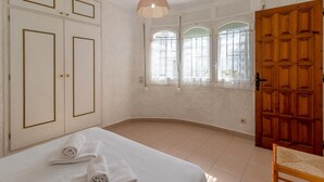 Un dormitorio con una cama, dos toallas enrolladas, una silla de madera, una puerta de madera y una ventana con barrotes de hierro decorativos.