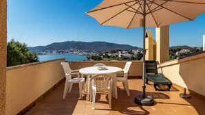 Una terrassa amb taula i cadires, una ombra i vista a una vila costanera i muntanyes.