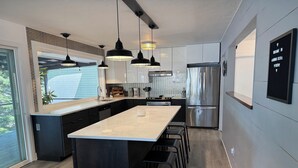 Cozinha moderna com ilha central, armários pretos, eletrodomésticos de aço inoxidável, iluminação pendente e vista através de uma grande janela.