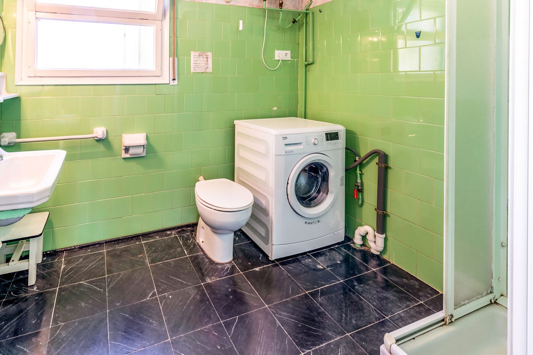 A small, compact bathroom with green walls, a black tiled floor, a white toilet, a washing machine, and a sink.