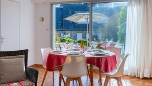 A dining area with a table set for four, large windows with curtains, and a view of a building and greenery outside. - La Sauvagère by Interhome (Carnac)