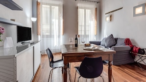A modern living room with a wooden dining table, black chairs, a grey sofa, a television, and a wine bottle.