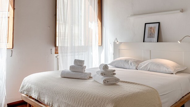 A bedroom with a bed, rolled towels, a framed picture, and a window with sheer curtains.