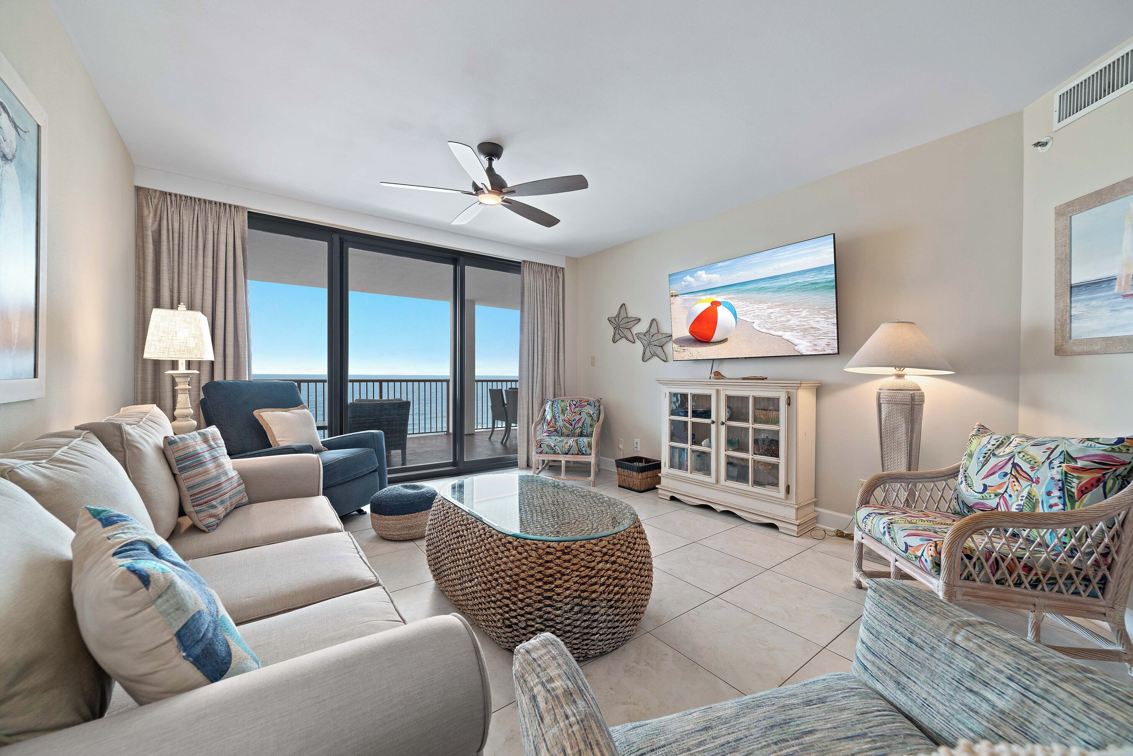 A spacious living area with a ceiling fan, a large flat-screen TV, and a view of the ocean through large windows.