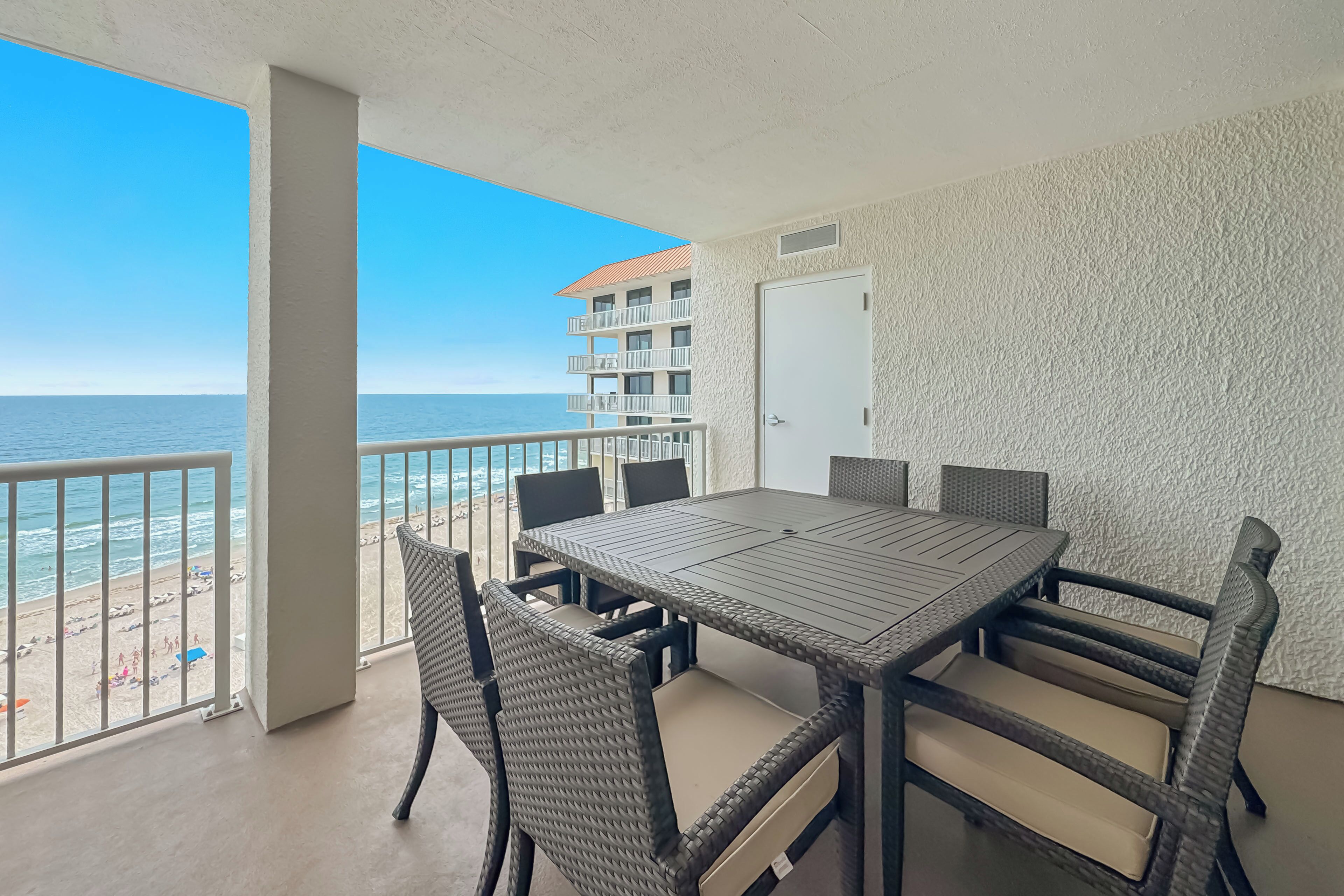 Un balcón con vista a la playa y al océano, una mesa de comedor con sillas, y un edificio con balcones al fondo.