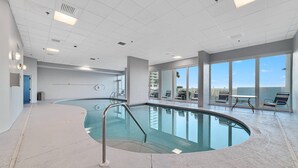 Une vaste piscine intérieure à la forme incurvée, entourée d’un sol carrelé et de grandes fenêtres offrant une vue sur la ville.