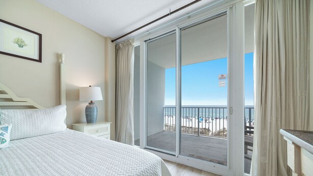 Une chambre à coucher comprenant un lit, une table de chevet, une lampe, un tableau encadré et une porte-fenêtre coulissante donnant sur un balcon avec vue sur la plage.