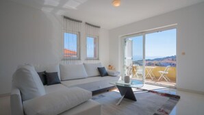 Un salon moderne avec un canapé blanc, une table basse en verre et une vue sur un balcon aménagé avec des sièges d’extérieur.