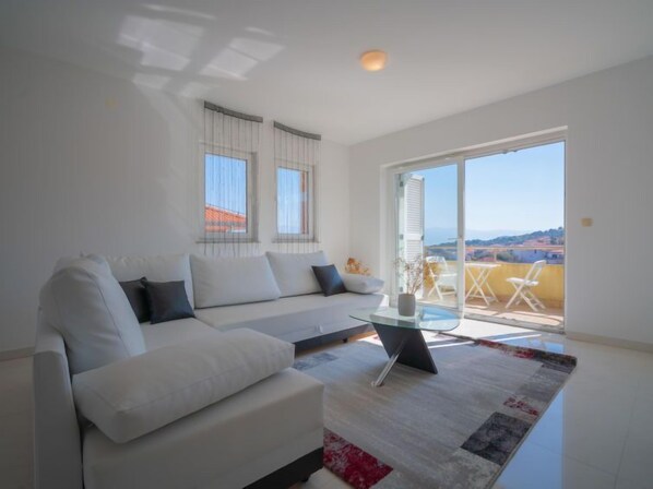 A modern living room with a white sofa, a glass coffee table, and a view of a balcony with outdoor seating.