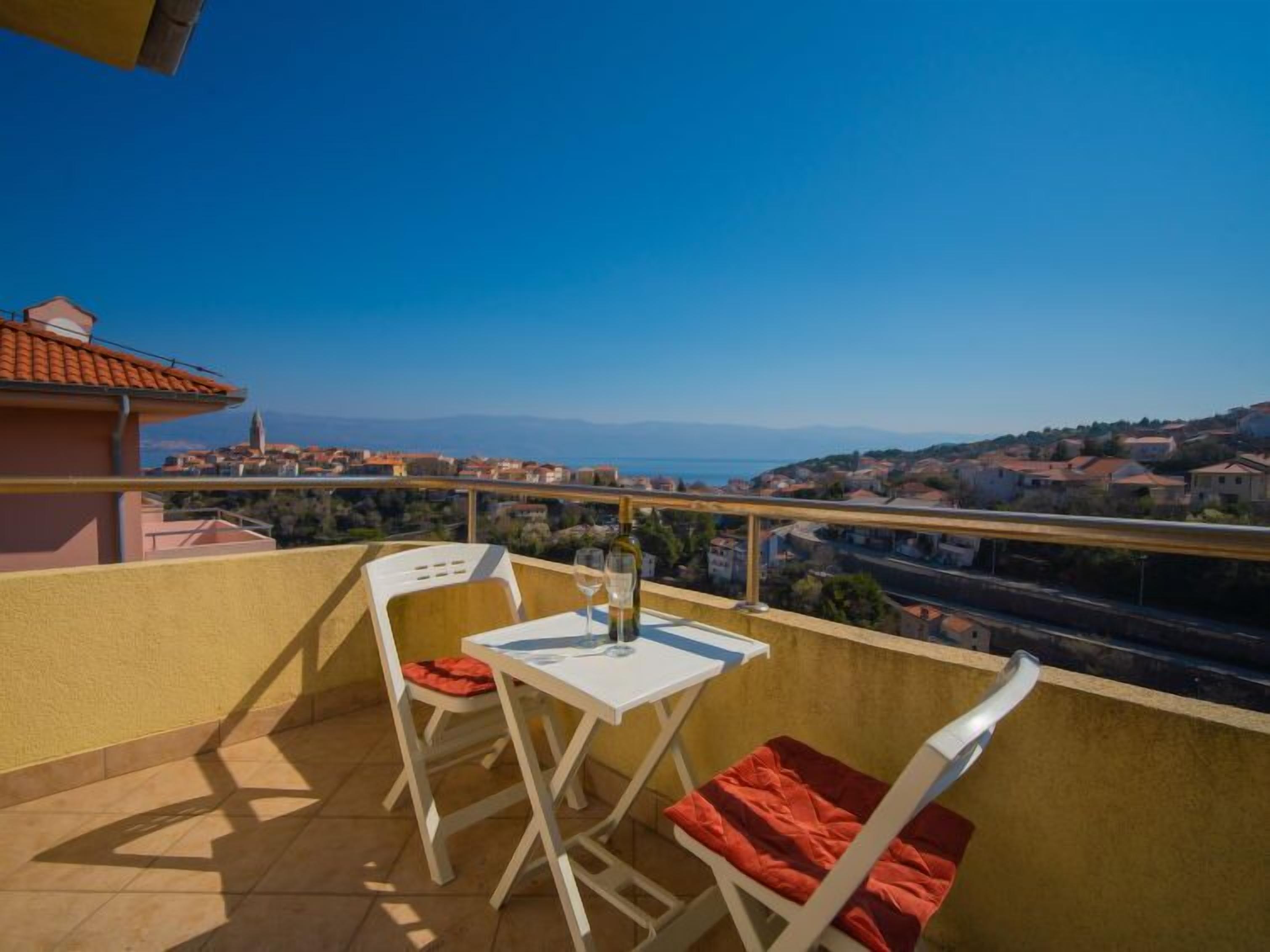 A balcony with two white chairs and a table, overlooking a coastal town with red-roofed buildings.