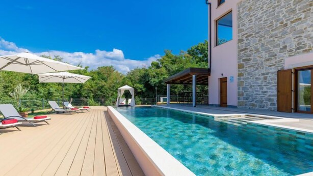 Un área de piscina moderna con sillones de descanso y sombrillas, una pared de piedra y un cielo azul despejado.