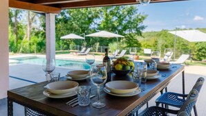 Un área de comedor al aire libre con una mesa de madera dispuesta para una comida, con vista a una piscina y abundante vegetación.