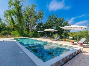 A swimming pool with tiled edges, surrounded by lounge chairs and umbrellas, set against a backdrop of trees and clear sky. - Villa Julia by Interhome (Buje)