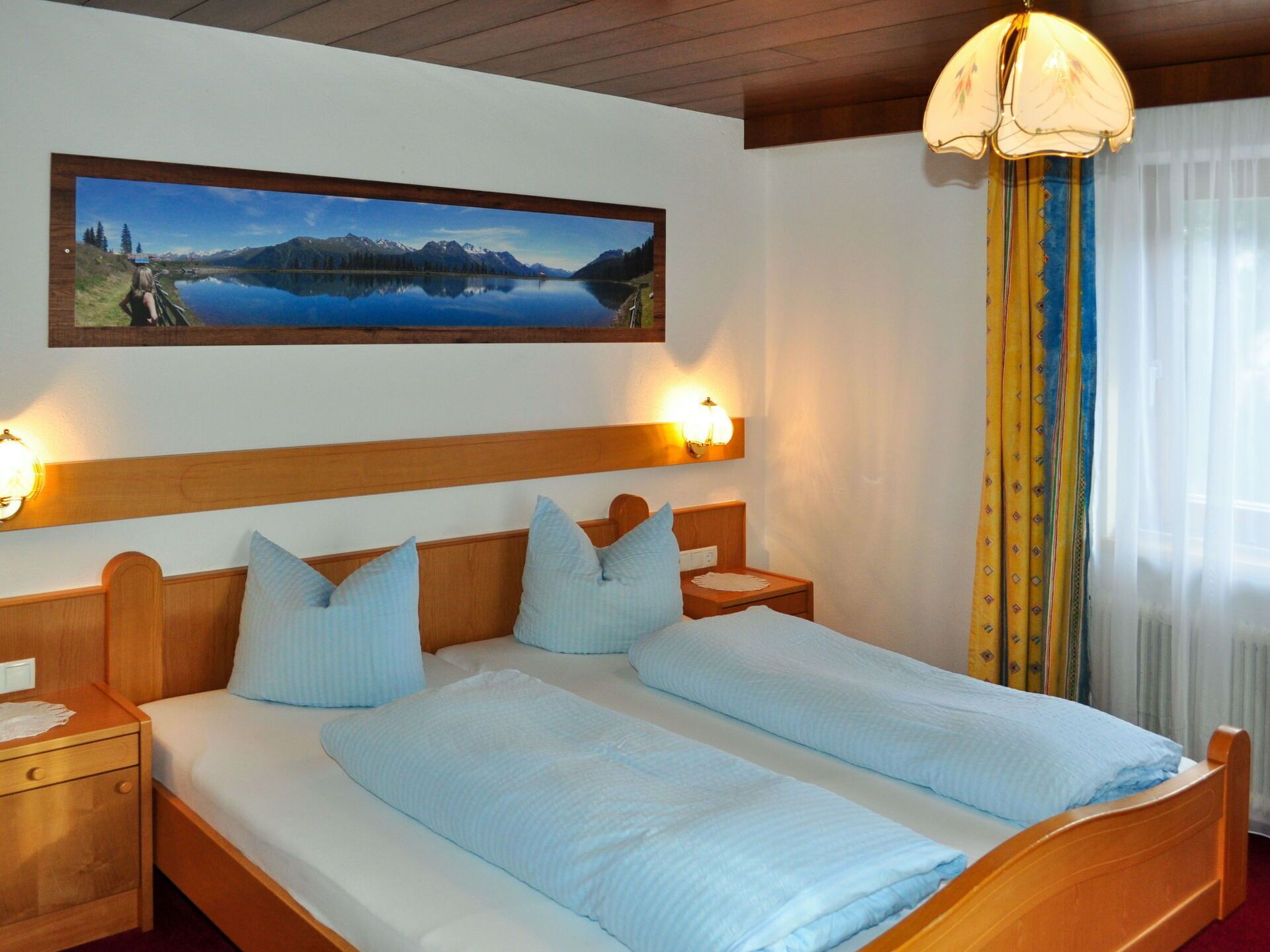 Cama doble con ropa de cama azul claro, cabecera de madera, dos mesitas de noche con lámparas, un cuadro de un lago y una ventana con cortinas.