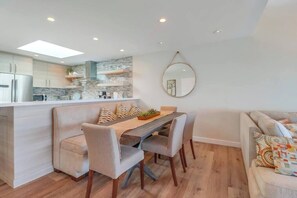 A modern kitchen with a central island, a dining area with a wooden table and chairs, and a living area with a sofa and cushions.
