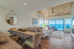 Ein großzügiger Wohnbereich mit Esstisch aus Holz, einer Couch und einem großen Fenster mit Blick auf das Meer.
