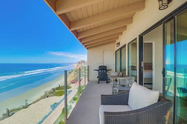 A balcony with a view of the beach and ocean, a grill, and a wicker sofa.