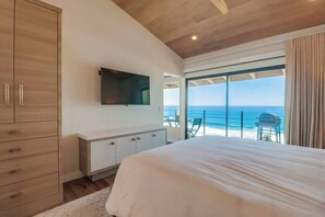 Ein Schlafzimmer mit einem großen Bett, einem an der Wand montierten Fernseher, einem Holzschrank und Blick auf den Strand durch große Fenster.