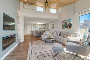 A spacious living room with a large sofa, a coffee table, and a fireplace. There is a kitchen in the background with modern appliances and a dining area.