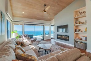 A spacious living room with a large sofa, a coffee table, and a fireplace. There is a television mounted on the wall and a ceiling fan. The room has large windows with a view of the ocean. - Oceanfront Luxury Condo w/ Hot Tub & Pool (Solana Beach)