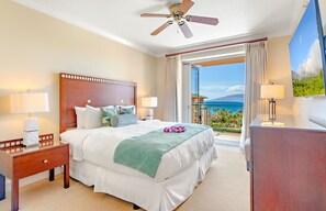 A hotel room with a bed, a nightstand, a desk, a ceiling fan, and a large window with a view of the ocean.