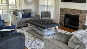Un salon avec un canapé gris, un fauteuil assorti, une table basse capitonnée, une cheminée et un ventilateur de plafond.