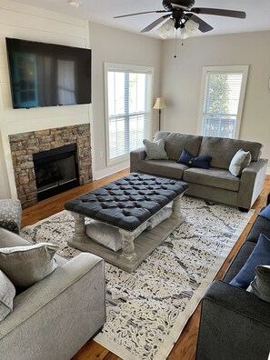 A living room with a stone fireplace, a grey sofa, a tufted ottoman, and a ceiling fan.