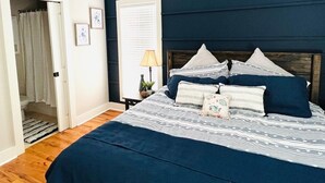 Une chambre avec un grand lit, un ventilateur de plafond, une fenêtre avec des stores et une porte donnant sur une salle de bain.