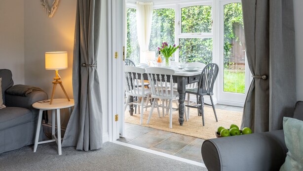 Un salon chaleureux avec un canapé gris, une petite table d’appoint avec une lampe, et un coin repas comprenant une table et des chaises.