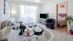 A modern, well-lit room with a dining table set for two, a TV, a comfortable armchair, and a bed with a patterned bedspread. - L'Hippocampe T2 avec Balcon by Interhome (Les Sables d'Olonne)
