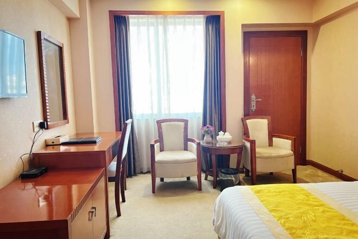 Une chambre d’hôtel comprenant un lit, un bureau, deux chaises, une petite table avec une théière et une fenêtre avec des rideaux.