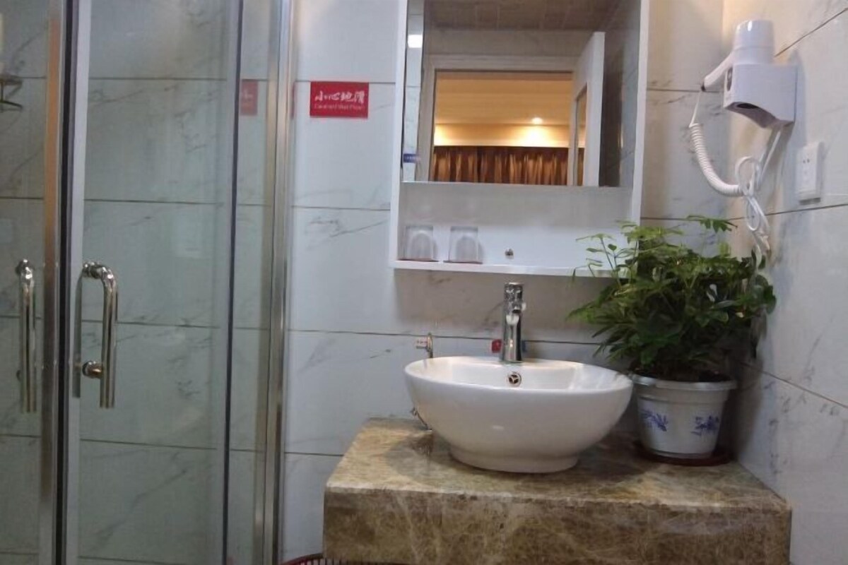 Une salle de bain dotée d’une douche à parois de verre, d’un lavabo blanc avec un plan de toilette en marbre, d’une plante en pot et d’un sèche-cheveux fixé au mur.