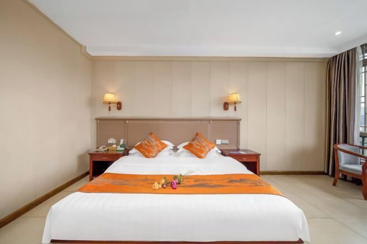 A hotel room with a large bed, two bedside tables, a nightstand, a chair, and a window with curtains.
