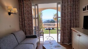 A modern hotel room with a grey sofa, a small white table with wine glasses, a TV on a cabinet, and a balcony with a view of the sea. - Le Village (E1 121) by Interhome (Agay Village Cap Esterel)
