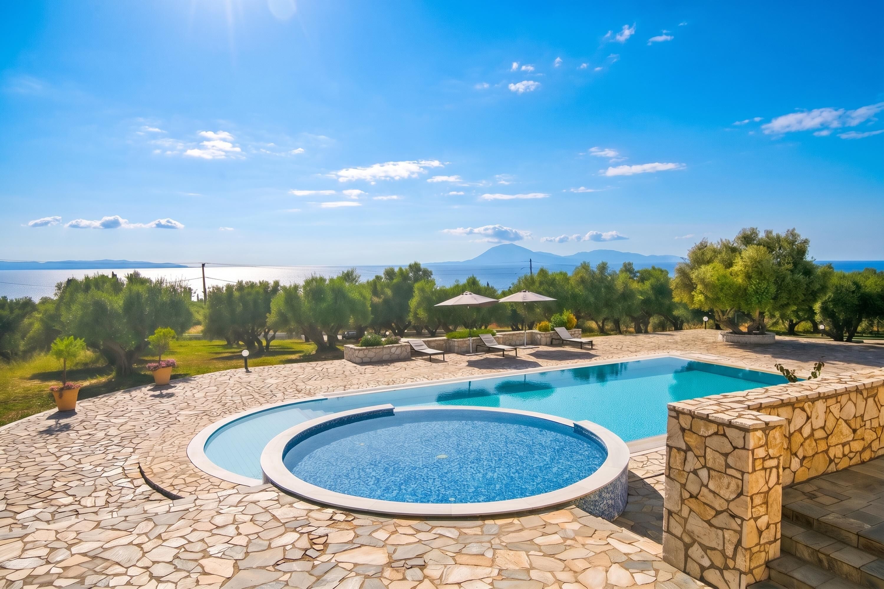 A swimming pool with a stone border, surrounded by a stone patio, and overlooking a body of water with mountains in the distance.