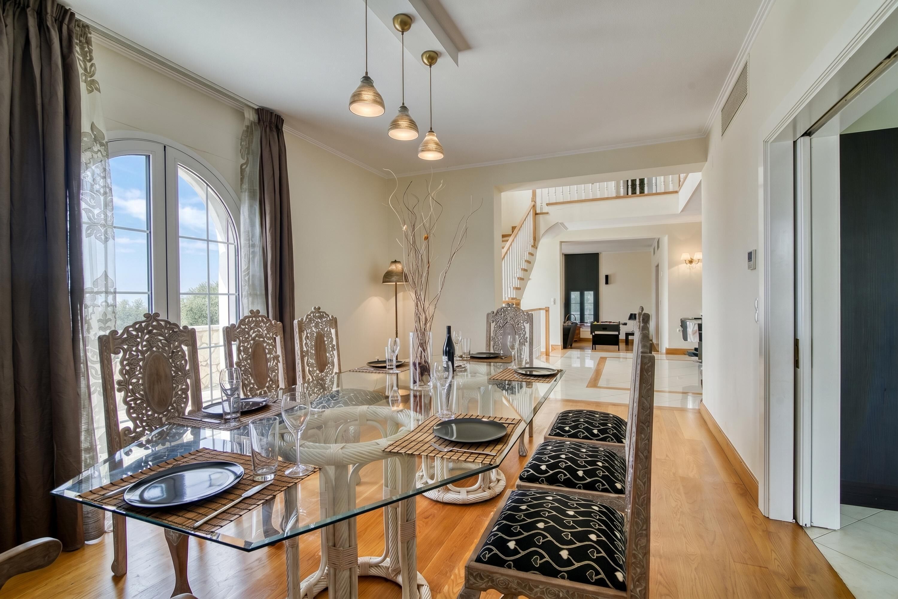 A spacious dining area with a glass table, ornate chairs, and a staircase in the background.