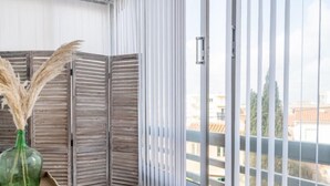 Un balcón con vistas a edificios, una barandilla de madera con listones horizontales, un jarrón verde con plantas secas y una silla de mimbre.