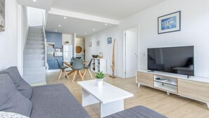 A modern living room with a grey sofa, a white coffee table, and a TV on a wooden TV stand. There is a staircase leading to a kitchen and dining area. - Port Canigó by Interhome (Roses)