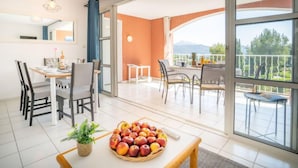 Een lichte kamer met een eettafel, stoelen en een fruitschaal. Er is een groot raam met uitzicht op groen en bergen.