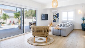Ein modernes Wohnzimmer mit einem großen Glasfenster, das zu einem Außenbereich führt, einem grauen Sofa, einem Korbstuhl und einem runden Teppich.