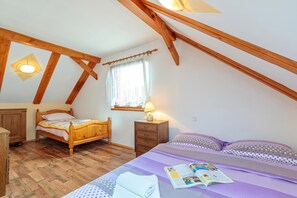 Een slaapkamer met een schuine kap, houten balken, een bed met een gebedrukte sprei, een nachtkastje met een lamp en een raam met gordijnen.