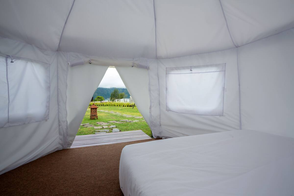 Ein Zeltraum mit Bett, Fenster und Blick auf eine Wiese mit einem Kiesweg und einem Gebäude in der Ferne.