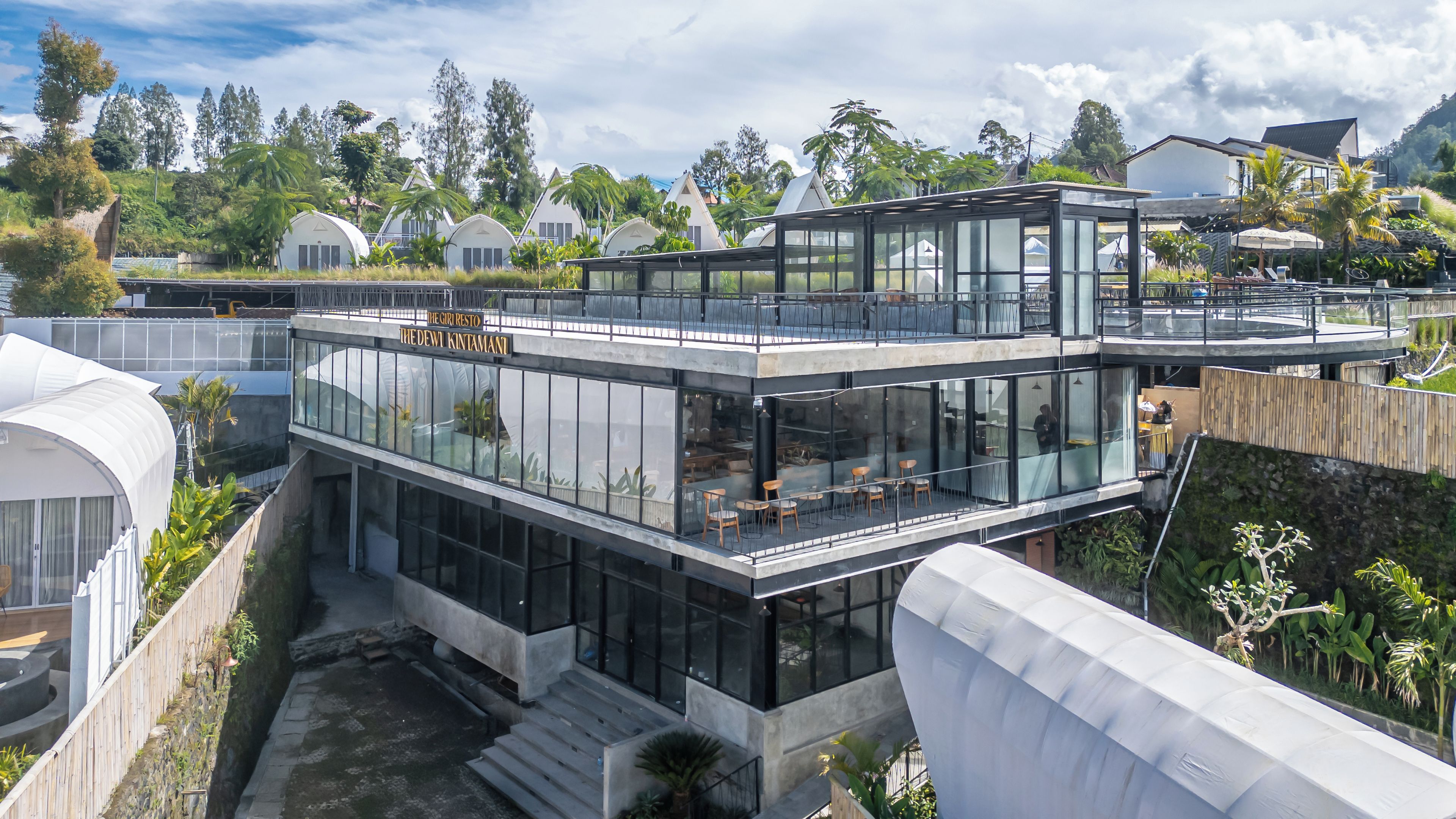 Un edificio moderno de varios pisos con fachadas de vidrio, mesas al aire libre y un letrero que indica 'The Giri Resto'.