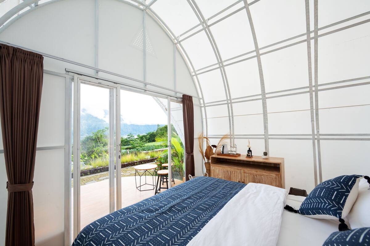 Ein Schlafzimmer mit einem Bett, einem Nachttisch aus Holz und Blick auf einen Garten durch große Fenster.