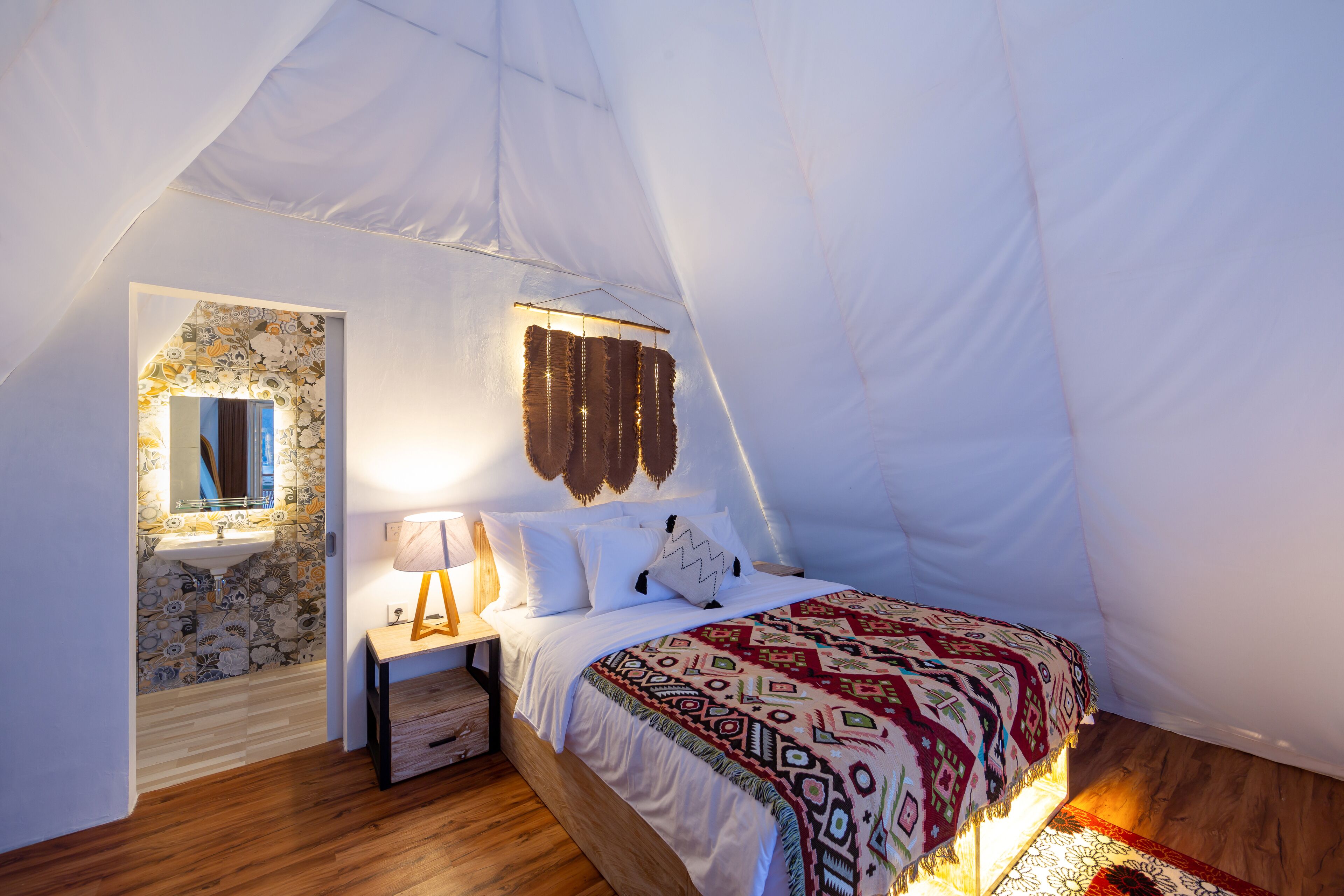 A tent-style bedroom with a bed, bedside table, lamp, and a small bathroom with a mirror and sink.