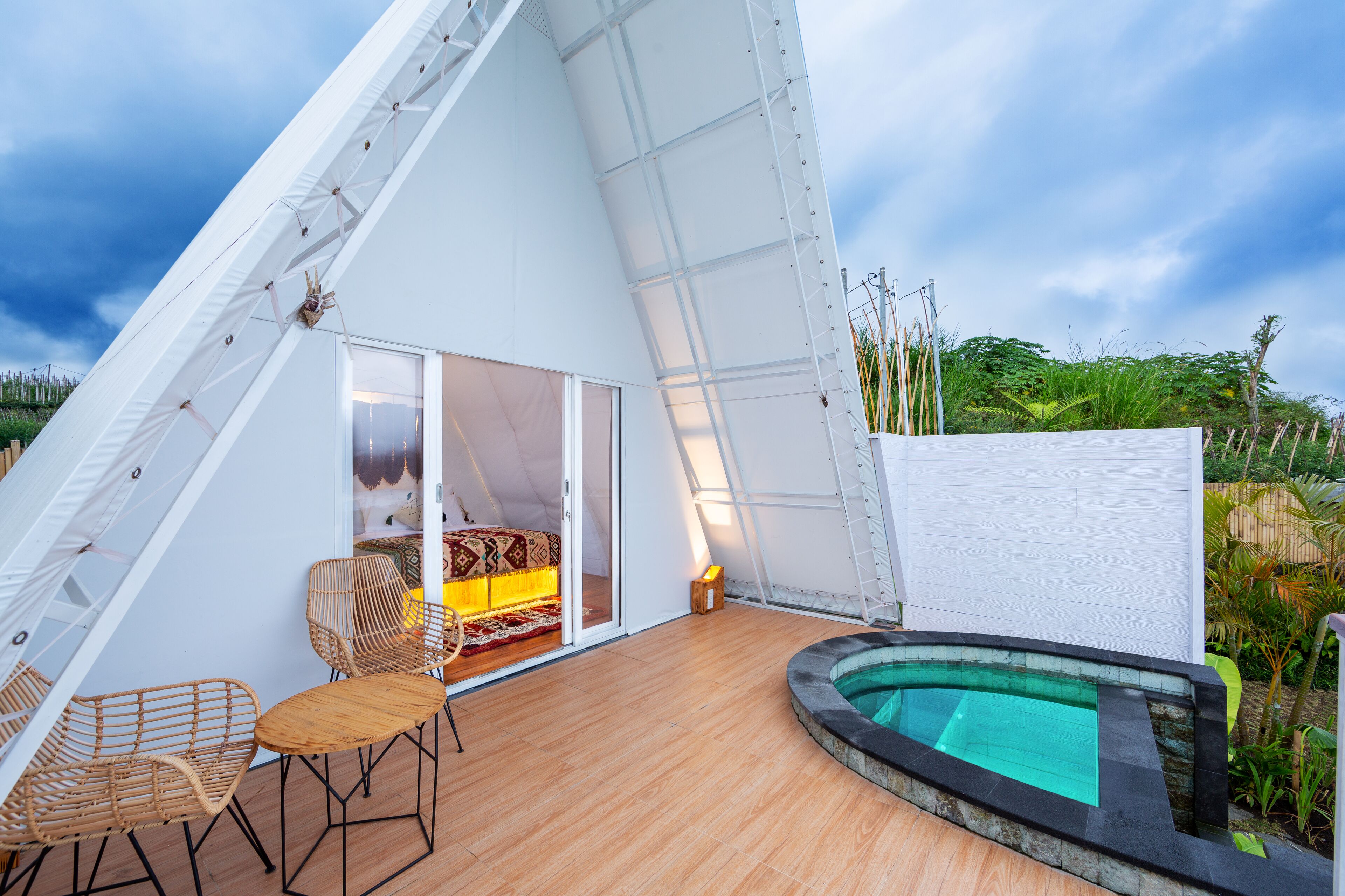 Una estructura moderna, de color blanco y techo triangular, con piscina, piso de madera y mobiliario de mimbre.