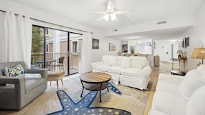Un salon spacieux avec un canapé blanc, un fauteuil gris et une table basse en bois. On y trouve un ventilateur de plafond, une grande fenêtre avec des rideaux et un balcon agrémenté de mobilier d’extérieur.