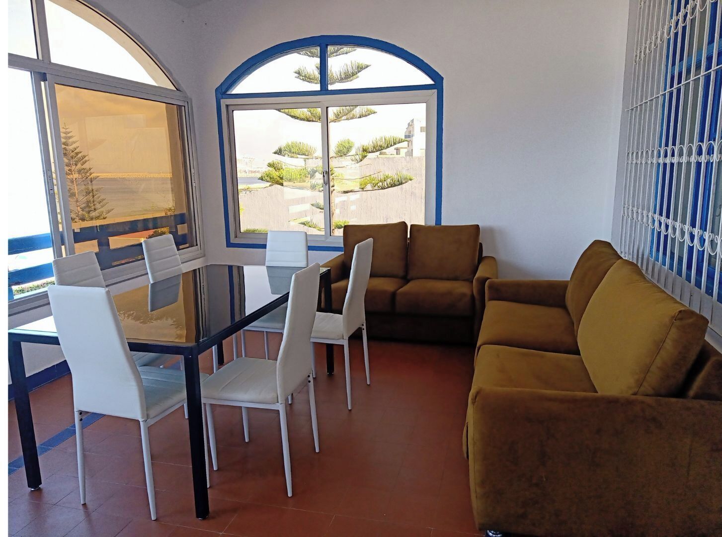 Ein Raum mit einem Glastisch, umgeben von Stühlen, einem braunen Sofa und einem großen Fenster mit Blick auf Grünflächen und Gebäude.